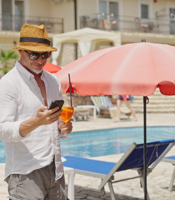 Male tourist using smartphone and holding cocktail by hotel pool on sunny summer day. Middle aged man on vacation reading email or checking social media on cell phone.